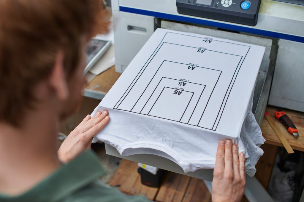 High angle view of young blurred craftsman working with t-shirt with marking and screen printing